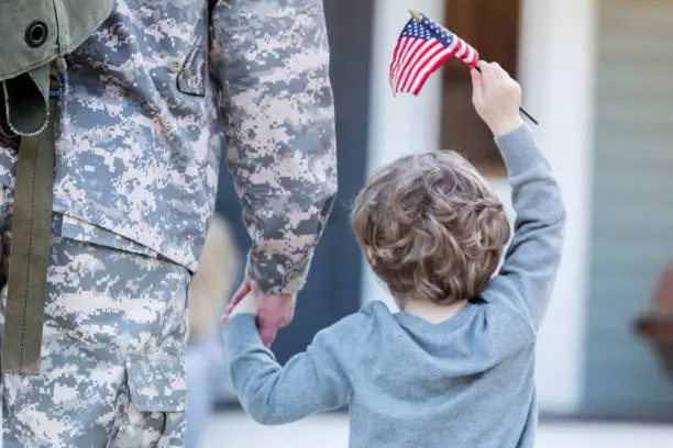 Rear view of preschool age boy holding his army dad's hand. They are walking toward their home. The boy is holding an American flag.