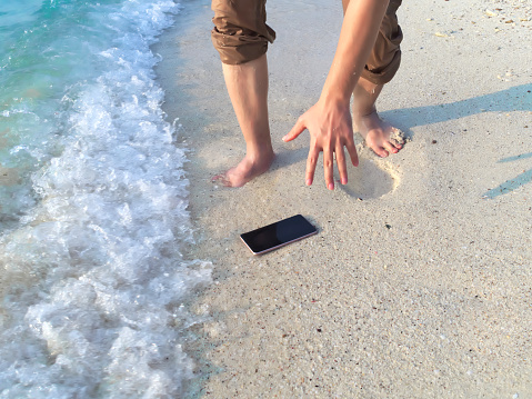 Hands of young Asian man dropping mobile smart phone on tropical sandy beach. Accident and insurance electronic equipment concept