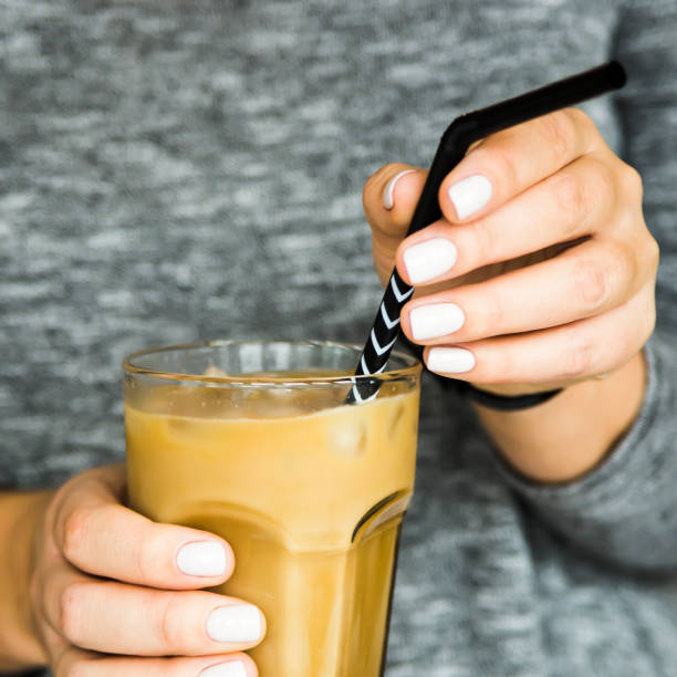A girl in a gray dress adjusts a black straw in a large glass with ice-cold coffee with milk. Summer cooling drink concept