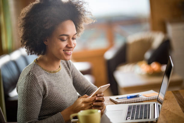 istock Happy black woman using smart phone while working on laptop at home. 932651362