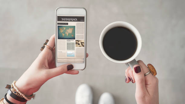 donna che tiene il cellulare e legge notizie dallo schermo altra mano che tiene la tazza di caffè - newspaper reading holding paper foto e immagini stock