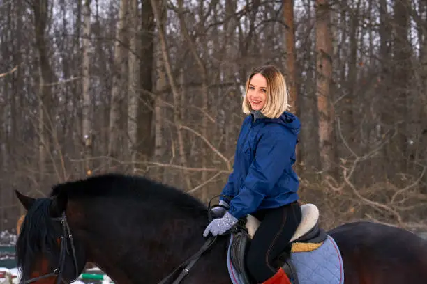 A young girl with white hair learns to ride a horse. The girl recently started to practice equestrianism. The girl is afraid of riding a horse quickly. A cloudy winter day."n
