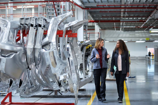 Engineers walking while discussing by car chassis Female engineers discussing by car chassis. Full length of professionals are walking on aisle in factory. Colleagues are in automobile industry. production line automobile industry car plant car stock pictures, royalty-free photos & images