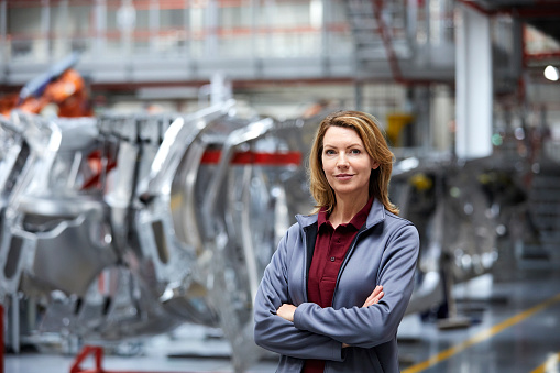 Portrait of confident engineer with arms crossed by car chassis. Young female professional is standing in car plant. She is working in automobile industry.
