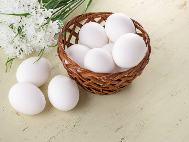 huevos frescos en una cesta y sobre una mesa de madera. contra el fondo de flores blancas - animal egg eggs basket yellow fotografías e imágenes de stock