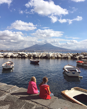 Two young girls sitting on the harbour dock looking out across the Bay of Naples to Mount Vesuvius volcano