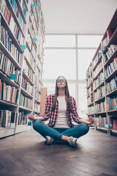 tiro de ângulo baixo de jovem garota nerd calma, praticando yoga em posição de lótus no chão de madeira na sala de arquivo da biblioteca da faculdade - balance book university glasses - fotografias e filmes do acervo