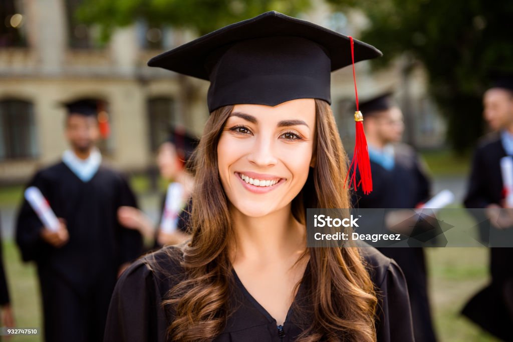 Feliz chica linda morena grad caucásico es sonriente, sus compañeros de clase están detrás. Ella está en una Junta de mortero negro, la borla roja en el vestido, con pelo rizado marrón agradable. ¡Grado académico de maestro! ¡Un éxito! - Foto de stock de Graduación libre de derechos