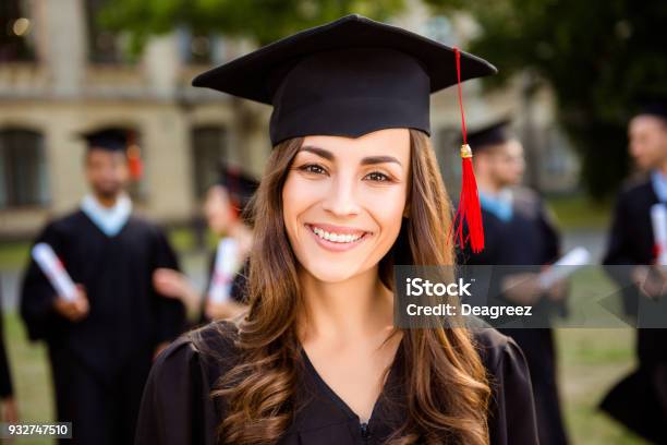 Feliz Chica Linda Morena Grad Caucásico Es Sonriente Sus Compañeros De Clase Están Detrás Ella Está En Una Junta De Mortero Negro La Borla Roja En El Vestido Con Pelo Rizado Marrón Agradable Grado Académico De Maestro Un Éxito Foto de stock y más banco de imágenes de Graduación