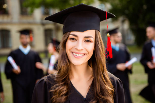 happy cute brunette caucasian grad girl is smiling, her class mates are behind. she is in a black mortar board, with red tassel, in gown, with nice brown curly hair. master degree! success! - graduation student women beauty imagens e fotografias de stock