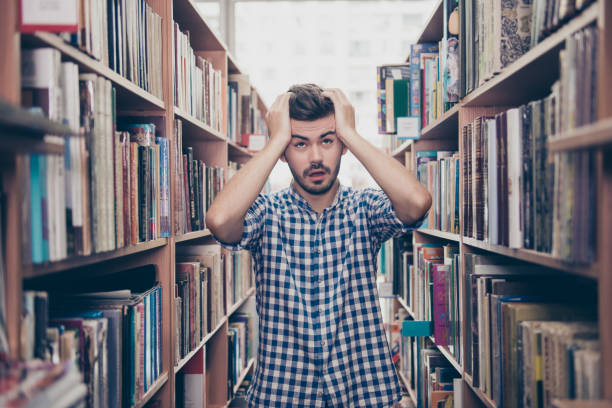 istock Stressed young brunet bearded student`s brain is blasting, he stands between the book shelves in the library, tired shocked grimace, holding the head, too much information! Exam is soon 932750602