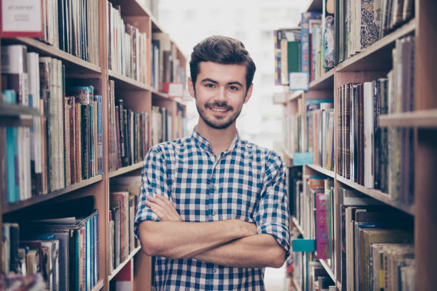 istock Cheerful clever young attractive brunet bearded student bookworm is standing with crossed hands in the ancient university library, between book shelves, smiling, wearing casual checkered shirt 932750836