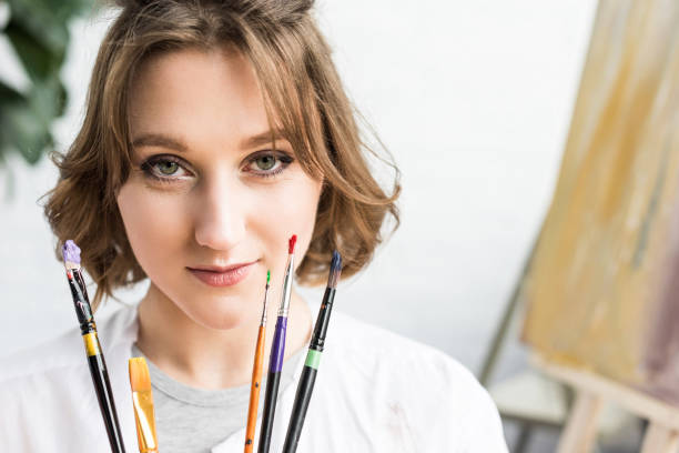 istock Young inspired girl looking at camera with brushes in hands in light studio 932760750