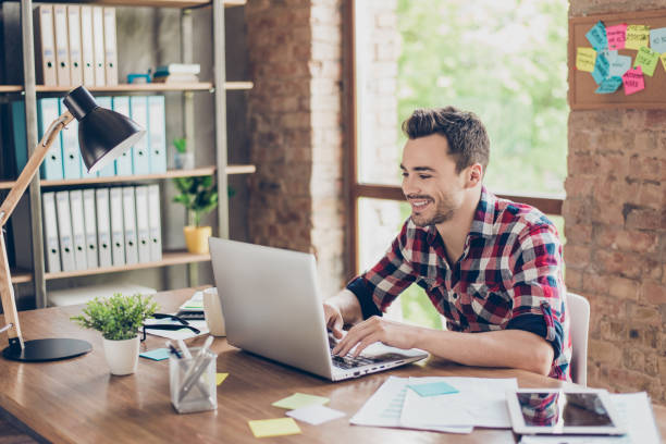 istock Cheerful young brunet freelancer is smiling, typing on his laptop in nice modern work station at home, in casual smart wear 932763106
