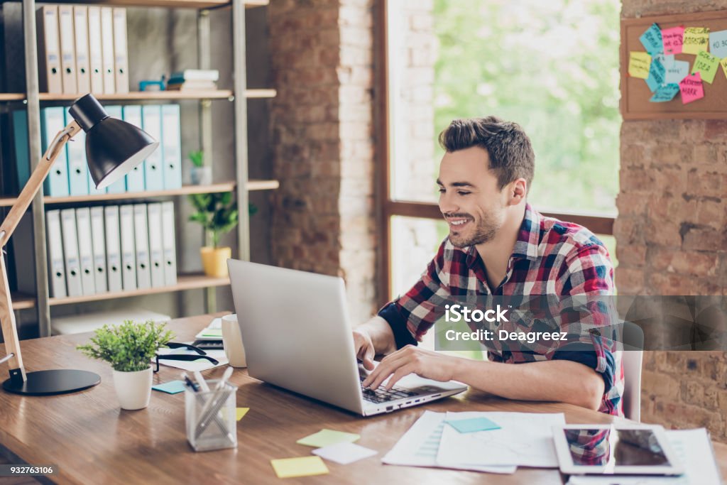 Cheerful young brunet freelancer is smiling, typing on his laptop in nice modern work station at home, in casual smart wear Laptop Stock Photo