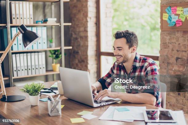 Cheerful Young Brunet Freelancer Is Smiling Typing On His Laptop In Nice Modern Work Station At Home In Casual Smart Wear Stock Photo - Download Image Now