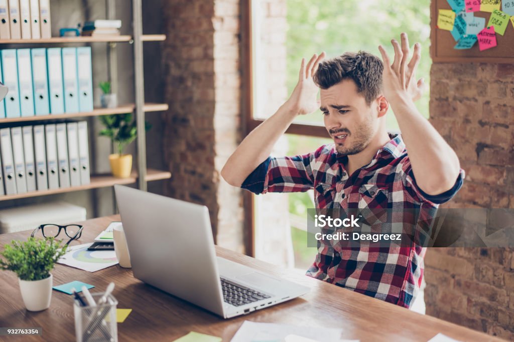 Wtf?! Omg! Oh no! Young amazed emotional brunet with bristle with sad grimace looks at screen of device, he can`t believe the bad news he received, sitting at workstation, gesturing with palms of arms Distraught Stock Photo