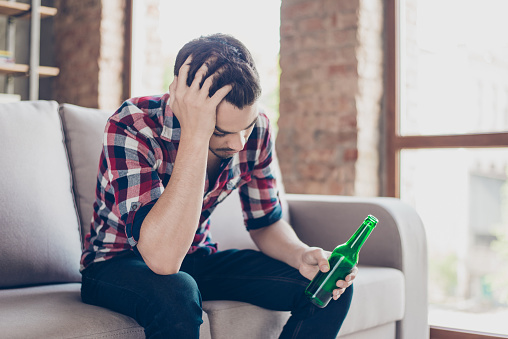 Bad luck, alcohol addiction, relationship problems. Frustrated young guy with bristle is holding his head, he is upset because his business has failed. He is having beer and sitting on the couch alone