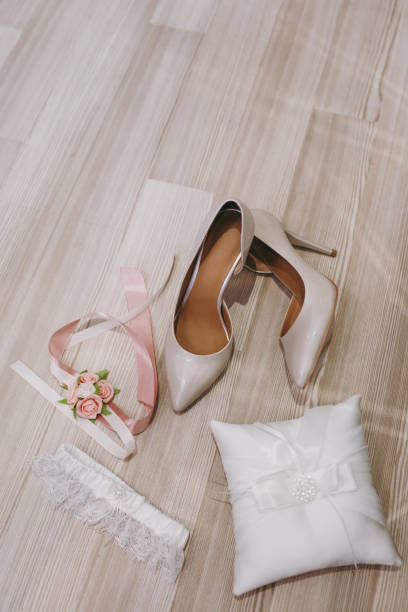 istock Close up Elegant light beige wedding varnish shoes with sharp noses, pillow for rings, boutonniere with pink flowers and garter for the bride on a wooden floor background. Wedding wear, accessories 932768880