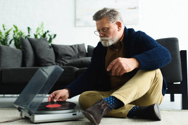 istock stylish senior man in eyeglasses putting vinyl record in turntable while sitting on carpet 932779040