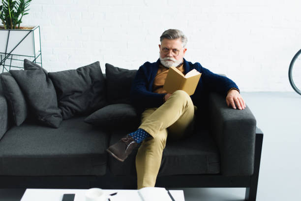 istock stylish bearded man in eyeglasses holding book and looking at camera while sitting on sofa at home 932779198