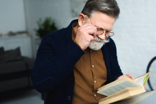 istock smiling bearded senior man in eyeglasses reading book at home 932785580