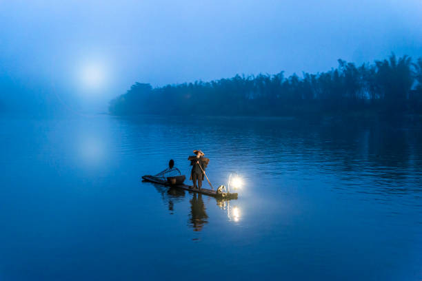 Traditional Chinese 75 year old senior fisherman in traditional clothes and bamboo hat on his wooden fishing raft with two cormorants fishing on the Li River in the early morning fog light at sunrise. Shot at Xing Ping, close to the city of Yangshuo County, Guangxi, Guilin, China. tradicional china 75 años senior pescador tradicional ropa y sombrero de bambú en su balsa de pesca de madera con dos cormoranes pescando en el río li en la niebla de la mañana luz al amanecer. disparo en xing ping, cerca de la ciudad de yangshuo cuenta