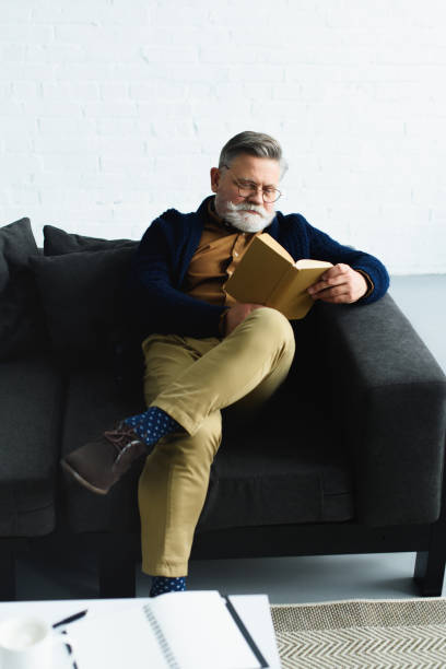 istock stylish bearded man reading book on sofa at home 932790062
