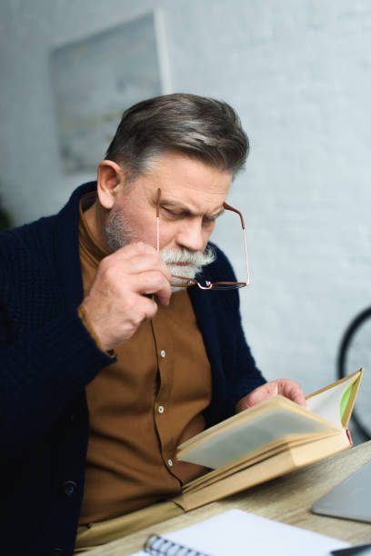istock bearded senior man adjusting eyeglasses and reading book at home 932790176