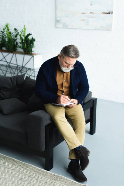 istock high angle view of bearded senior man sitting on sofa and writing in notebook 932790318