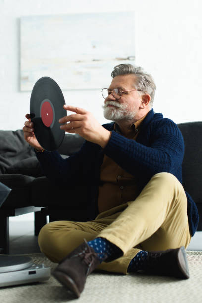 istock smiling stylish senior man in eyeglasses holding vinyl record while sitting on carpet 932790486