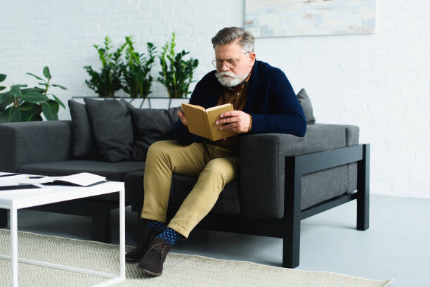 istock serious bearded senior man sitting on sofa and reading book at home 932790696