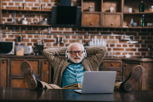 istock Relaxed old man in headphones using laptop with feet on table 932841630