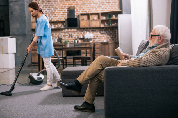 istock Senior man reading while caregiver vacuums room 932845018