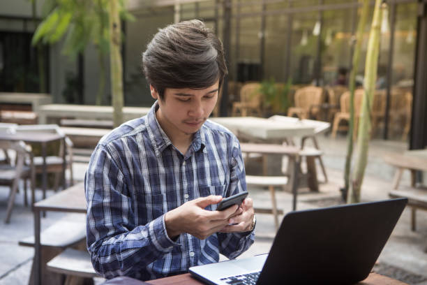 istock Southeast asian young man using his smartphone and laptop computer at the outdoor cafe 932846340
