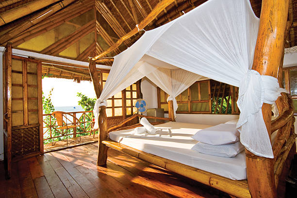 A series of images showcasing a rustic-style wooden beach cabana with a thatched roof and flowing white curtains, located on a sunny Mediterranean or tropical shoreline. The cabana is furnished with soft beige cushions and a wooden table, offering a serene and private space for relaxation. The interiors open up to a tranquil beach scene with sun loungers, parasols, and clear blue waters in the background. The light breeze moves the curtains gently, creating a peaceful, idyllic vacation atmosphere perfect for themes of luxury travel, summer leisure, and eco-friendly beachfront architecture.