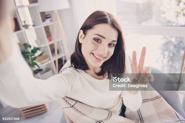 Self Portrait Of Pretty Charming Nice Cute Stylish Brunette Girl In Pullover With Modern Hairstyle Shooting Selfie Gesturing Vsign To The Front Camera Wrapped In Plaid Sitting In Living Room Stock Photo - Download Image Now