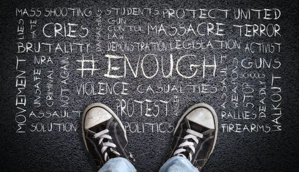 A teenager in jeans and canvas shoes standing on asphalt road with # ENOUGH word cloud. Concept of social movement to protest gun violence and mass school shootings in the United States.