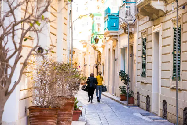 Color image depicting two women walking on a bright sunny, narrow street in the town of Mdina (also known as Rabat) in Malta. The quaint old houses have decorative plants outside. The women are in rear view and are walking into the distance. No one else is around, giving a calm peaceful atmosphere. One of the women is wearing a bright yellow jacket. Lots of room for copy space.
