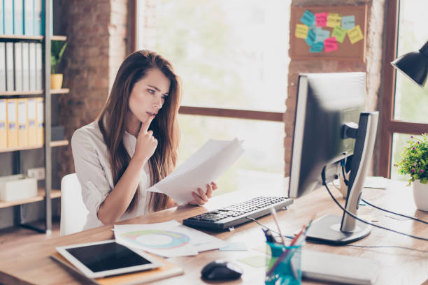 istock Beautiful pretty charming cute with modern hairstyle agent secretary trainee checking documents in hand looking at the monitor sitting at the table in light equipped with modern technology office 932959544