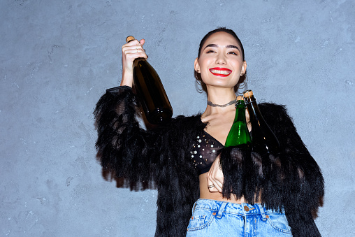 fashionable happy asian girl holding bottles with alcoholic beverages on grey