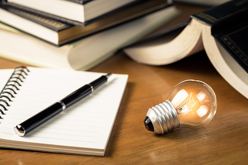 Small light bulb glowing on the desk, with notebook and many books on background, reading and writing idea concept