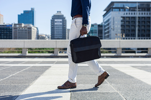 Mid adult African businessman walking through the city holding his laptop bag, the man is well dressed and the image is taken from the waist down.