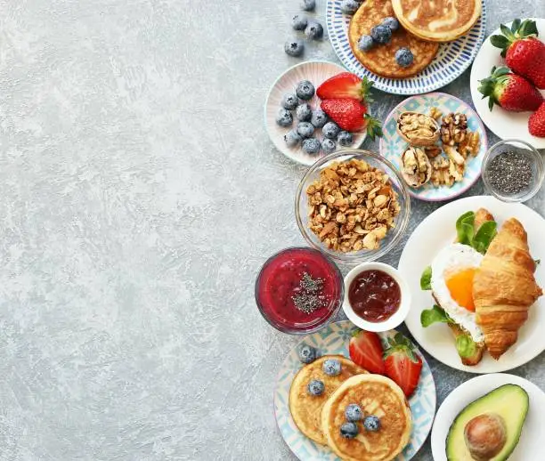 Breakfast food table. Festive brunch set, meal variety with granola, fried egg, pancakes, croissants, smoothie ,fresh vegetables, berries and fruits. Overhead view, copy space
