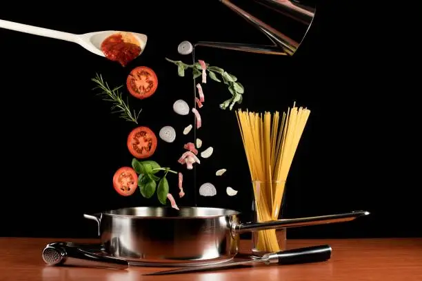Studio shot against a black background of lots of ingredients for spaghetti Bolognese. Almost all ingredients are falling from the top of the image into a large aluminium frying pan. Items like a knife, glass with pasta and garlic press are also displayed on the table.