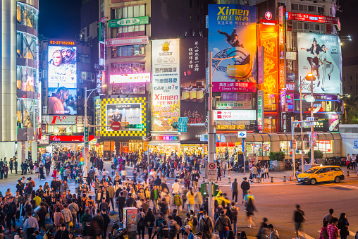 View across the crowded crossroads of Ximending past zooming traffic, shoppers and tourists enjoying the warm night illuminated by the colourful billboards and neon lights of Taipei’s downtown commercial district, Taiwan.