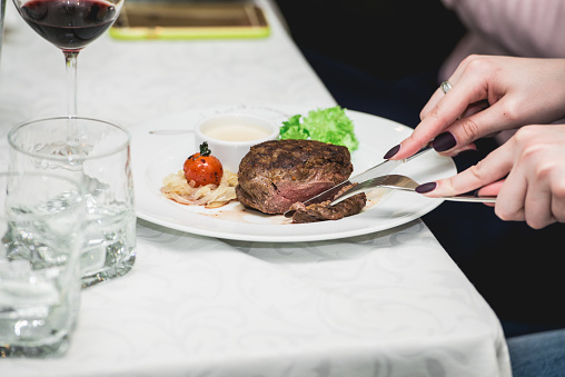 woman cuts a piece of fresh grilled bbq roast beef steak and sauce on a white plate with green leaf of salad. soup sauce small jug glass served on a table in a restaurant.