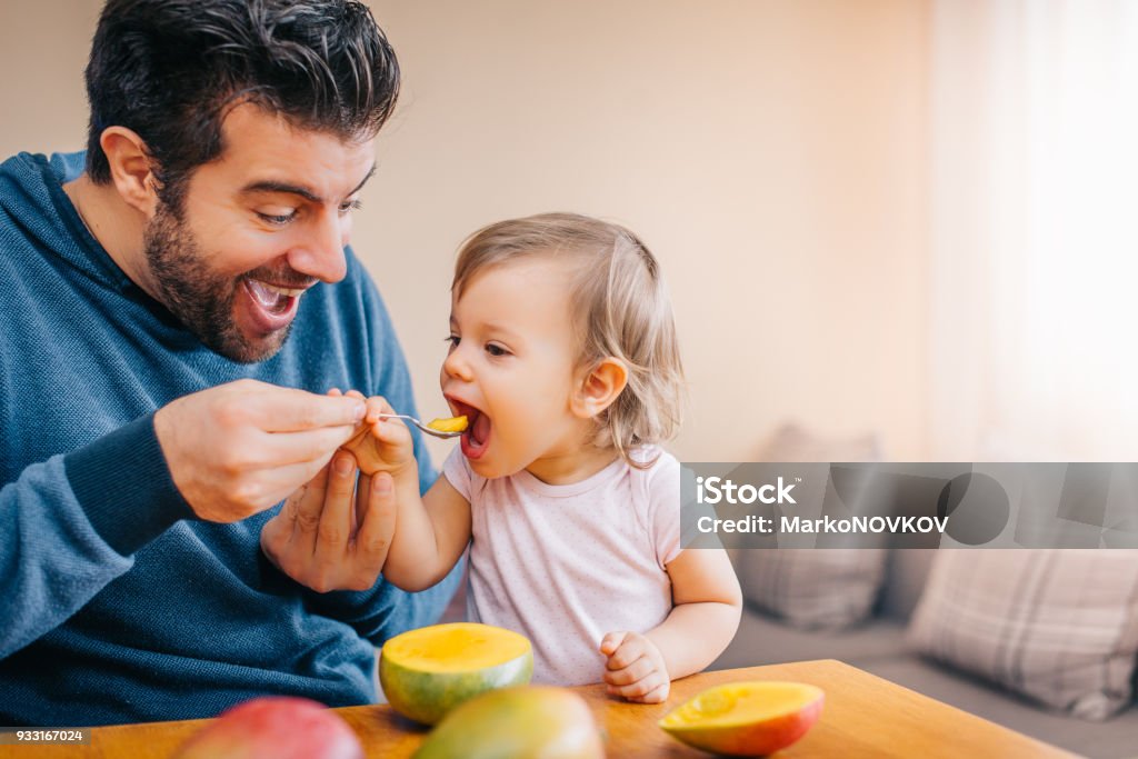 Père d’alimentation bébé bébé bambin avec cuillère et mangue - Photo de Bébé libre de droits