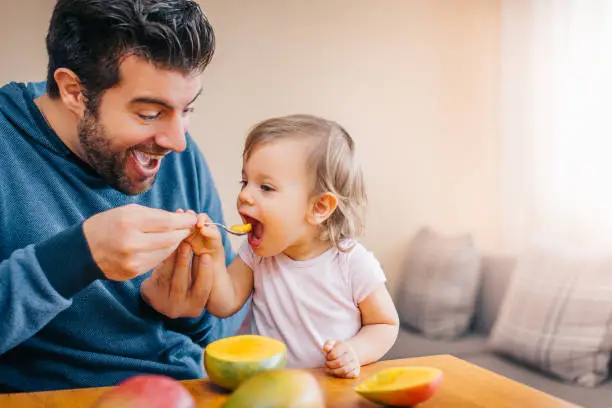 Photo of Father feeding toddler baby infant with spoon and mango