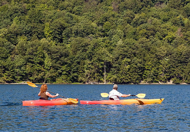 DSLR image of two mature adults kayaking Two mature adults, a man and a red headed woman, in kayaks on Canadice Lake, on of the Finger Lakes in New York State.  The bright sun is glistening on the rippled blue water, with green trees in the background.  The DSLR image is in horizontal orientation with ample copy space for text.   finger lakes stock pictures, royalty-free photos & images